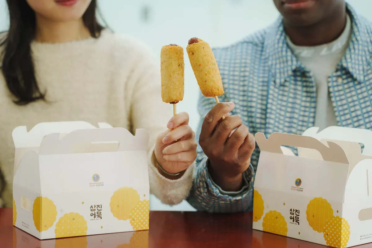 Two people holding skewered eomuk fish cakes in front of gift boxes from a Busan fish cake brand