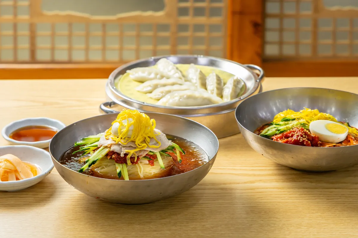 A metal bowl of milmyeon topped with sliced pork, cucumber, and egg, with steamed dumplings on the side