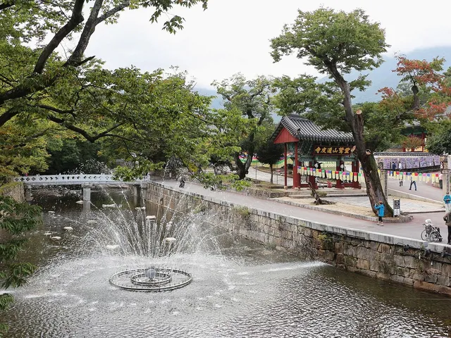 Pond and fountain at the Tongdosa entrance, a stone bridge, visitors walking and a red one-pillar gate bearing the sign 靈鷲山通度寺.