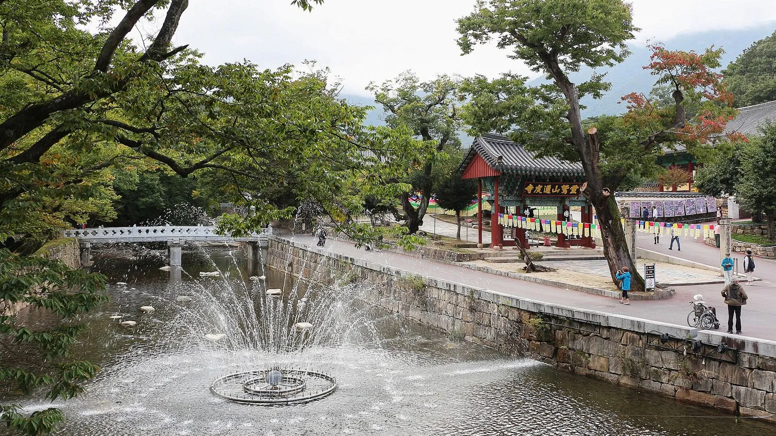 通度寺入口的池塘與噴泉、石橋，掛著「靈鷲山通度寺」匾額的紅色一柱門，以及步行的遊客。